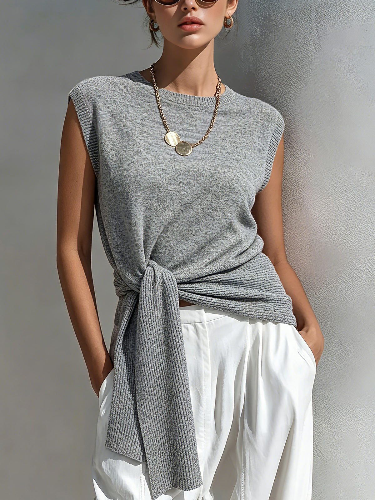 Woman wearing a gray sleeveless top with a knot detail and white pants against a neutral background