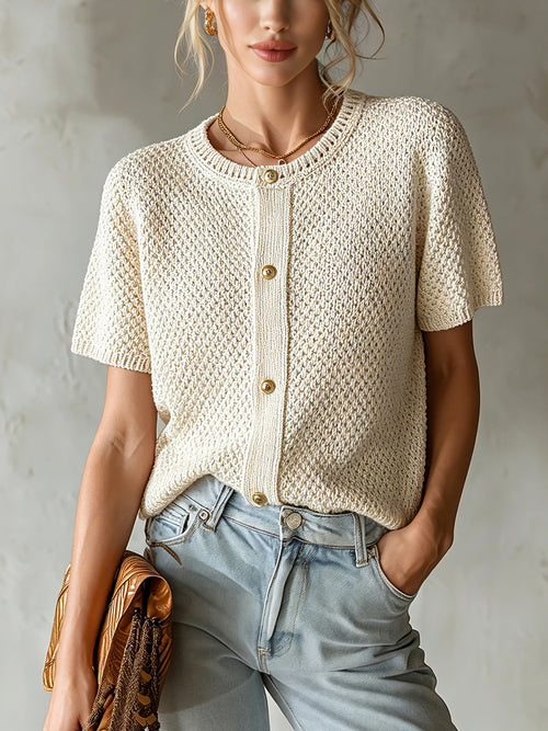 Woman wearing a beige knitted short-sleeve cardigan and light blue jeans against a neutral background